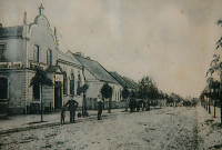 Historische Ansicht, eine von kleinen Linden umrahmte unbefestigte Strasse. Links im Vordergrund ein Gasthaus im Stil der Neigründerzeit.